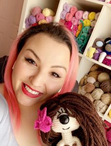 Mujer de cabello rosa sonriendo y sosteniendo una muñeca amigurumi ciclope con cabello cafe y flor en la cabeza